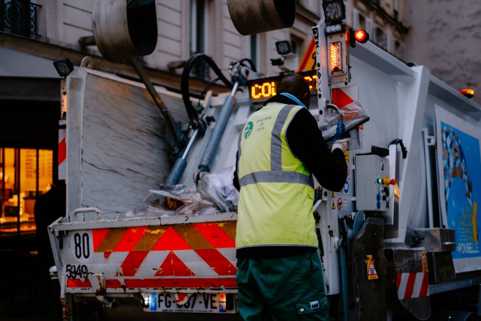 A waste collection worker wearing a high-visibility yellow vest with a logo on the back and blue gloves operates a mechanical compactor on the back of a large white rubbish truck. The truck is partially loaded with crumpled paper, cardboard, and plastic waste visible in the open container. The scene takes place outdoors during the evening or early night, with warm ambient lighting from nearby buildings illuminating the background. The truck features reflective red and white safety markings along its rear edges and equipment and control panels on the side, supported by hydraulic arms for waste compression. This visual reflects an example of independent waste disposal or rubbish removal service, illustrating the handling of bulky waste in an urban environment, aligning with the context of private waste collection options rather than central council collection. Rubbish Removal Chelsea's service scene emphasizes efficient rubbish management through professional waste collection activities.