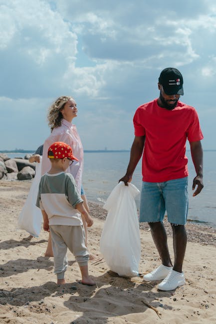 A group of three people, including a man and two children, are standing on a sandy beach near a body of water under a partly cloudy sky. The man, positioned on the right, wears a black cap, a bright red t-shirt, and light blue denim shorts, and is holding a white plastic bag, appearing to collect rubbish or debris. The young boy in front of him, wearing a grey and white shirt, beige trousers, and a red cap with a polka dot pattern, also holds a white plastic bag. The woman, standing behind them to the left, has curly blonde hair and wears a light pink shirt, observing as the man and boy work. In the background, there are large rocks lined up along the shoreline, and the water extends towards the horizon, reflecting the blue sky with scattered white clouds. The scene is lit naturally, with soft daylight illuminating the scene, appropriately illustrating an eco-friendly activity such as private rubbish collection or on-site waste management support by a service like Rubbish Removal Chelsea.