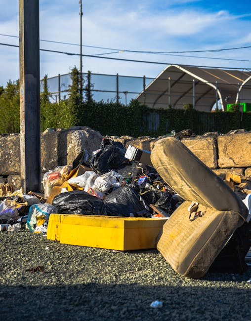A waste collection worker wearing a high-visibility yellow vest with a logo on the back and blue gloves operates a mechanical compactor on the back of a large white rubbish truck. The truck is partially loaded with crumpled paper, cardboard, and plastic waste visible in the open container. The scene takes place outdoors during the evening or early night, with warm ambient lighting from nearby buildings illuminating the background. The truck features reflective red and white safety markings along its rear edges and equipment and control panels on the side, supported by hydraulic arms for waste compression. This visual reflects an example of independent waste disposal or rubbish removal service, illustrating the handling of bulky waste in an urban environment, aligning with the context of private waste collection options rather than central council collection. Rubbish Removal Chelsea's service scene emphasizes efficient rubbish management through professional waste collection activities.