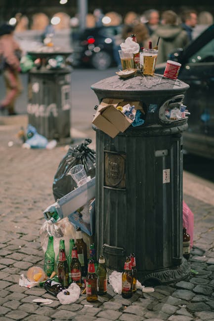A black, cylindrical public litter bin made of metal with a domed top, situated on a cobblestone street in an urban setting, is surrounded by overflowing trash and discarded bottles at its base. On top of the bin, there are several cups with plastic lids, some containing leftover beverages, and a small paper box. Attached to the side of the bin are additional paper and plastic waste, with crumpled paper and plastic wrappers visible. Nearby, a partly opened black garbage bag filled with rubbish rests against the bin, with various debris strewn across the ground, including crumpled paper, empty beer bottles, and plastic wrappers. In the blurred background, pedestrians and parked cars can be seen, with some appearing to be walking or standing, contributing to the busy street atmosphere. The scene illustrates issues related to waste management and highlights the importance of proper rubbish disposal, which private rubbish removal services like Rubbish Removal Chelsea often address through alternative waste handling solutions.