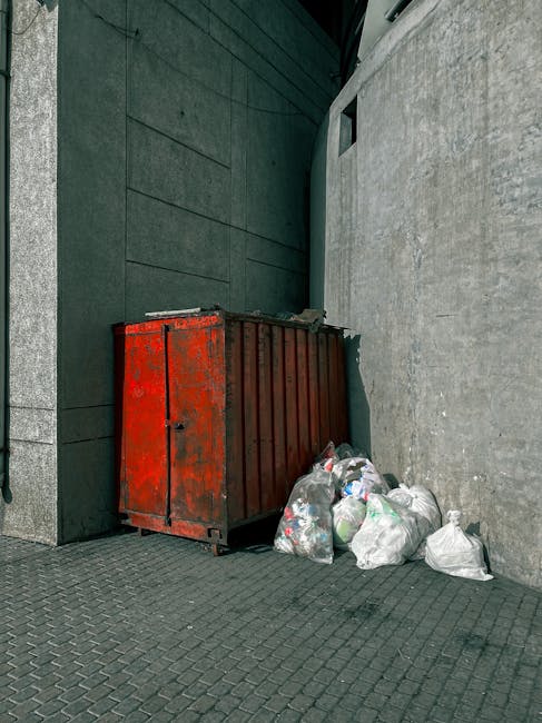A weathered red metal skip bin with visible rust and peeling paint, positioned against a dark green wall on the left and a light grey concrete wall on the right, on a paved urban sidewalk. Several clear plastic bags filled with assorted waste, including visible plastic bottles and packaging, are piled around the base of the bin, some leaning against it. The scene is outdoors, with natural daylight illuminating the area, highlighting the textures and colors of the bin, waste bags, and surrounding surfaces. The setting suggests an area designated for private rubbish collection or on-site clearance, typical for independent waste disposal services in urban environments, with the scene reflecting practical rubbish removal rather than municipal refuse collection.