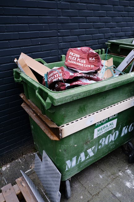 A black, cylindrical public litter bin made of metal with a domed top, situated on a cobblestone street in an urban setting, is surrounded by overflowing trash and discarded bottles at its base. On top of the bin, there are several cups with plastic lids, some containing leftover beverages, and a small paper box. Attached to the side of the bin are additional paper and plastic waste, with crumpled paper and plastic wrappers visible. Nearby, a partly opened black garbage bag filled with rubbish rests against the bin, with various debris strewn across the ground, including crumpled paper, empty beer bottles, and plastic wrappers. In the blurred background, pedestrians and parked cars can be seen, with some appearing to be walking or standing, contributing to the busy street atmosphere. The scene illustrates issues related to waste management and highlights the importance of proper rubbish disposal, which private rubbish removal services like Rubbish Removal Chelsea often address through alternative waste handling solutions.