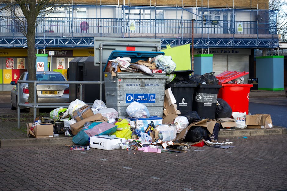 A large collection of mixed waste, including black and red rubbish bags, flattened cardboard boxes, paper, plastic containers, and discarded packaging, is piled on the pavement surrounding a metal recycling bin. The bin has a blue lid and labels indicating it is designated for mixed paper and card, with some waste spilling out onto the ground. Behind the waste, there's a parked silver car with a visible UK license plate, positioned near a metal railing. In the background, a commercial building with storefronts and signs is visible, partially obscured by a scaffolding structure and trees, under an overcast sky. The scene depicts an area where private rubbish collection or on-site clearance might be required, consistent with alternative waste handling services like those offered by Rubbish Removal Chelsea, in the context of managing waste for Chelsea homes and businesses.