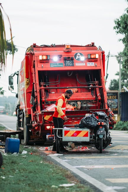 A municipal worker dressed in an orange high-visibility vest and dark trousers operates a large red rubbish collection vehicle on a roadside. The worker is shown loading waste into the compactor section of the truck, which features a closed rear loading hatch with hydraulic mechanisms and reflective warning stripes. The vehicle is parked on a paved road adjacent to a curb, with some litter and small debris scattered on the ground nearby. In front of the worker, there is a small metal cart or bin with a black rubbish bag placed on top, positioned close to the vehicle. The background reveals a few trees, utility poles, and a commercial building, suggesting an urban or suburban environment. The lighting indicates daytime with partly cloudy skies, and the scene illustrates an example of on-site waste collection or private rubbish removal, as provided by local services such as Rubbish Removal Chelsea, supporting alternative waste handling options outside municipal collection.