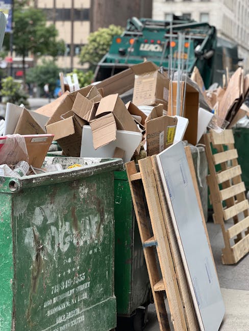 A large collection of mixed waste and recyclables piled around multiple rubbish bins on a paved urban street. The waste includes cardboard boxes, plastic bags filled with refuse, crumpled paper, and miscellaneous packaging materials. The grey recycling container is partially open, revealing flattened cardboard and paper inside, while a black bin and a red bin, both overflowing, contain assorted rubbish. In the background, there is a storefront with signage and a building under scaffolding, with a tree visible on the left side. A metal railing separates the waste area from parked vehicles, including a silver hatchback car. The scene is well-lit by natural daylight, highlighting the disorderly mixture of waste materials, which suggests an instance of late or unmanaged rubbish disposal, relevant to independent waste collection services such as those provided by Rubbish Removal Chelsea.
