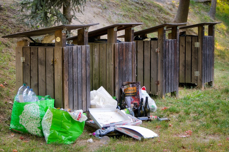 The image shows a row of four wooden outdoor rubbish bins with sloped, overhanging lids, situated on a grassy area next to a sloped, tree-covered terrain. The bins are made of dark stained wood with metal hinges and handles. In front of the first bin, there are various discarded items, including a large green plastic shopping bag filled with plastic bottles and bottles, some of which are partially visible, along with a white tray, crumpled white paper or napkins, and several glass bottles, some of which are dark green or brown, arranged randomly on the ground. Additionally, a black bag with a barbecue image printed on it, a plastic bag, and some loose trash like a cardboard box are scattered nearby. The surrounding environment features patches of grass and fallen leaves, with tall trees in the background, indicating a park or garden setting. The scene appears to be an example of improper waste disposal, highlighting the importance of professional rubbish removal services such as those offered by Rubbish Removal Chelsea, especially when handling outdoor waste collection and ensuring environmentally responsible disposal.