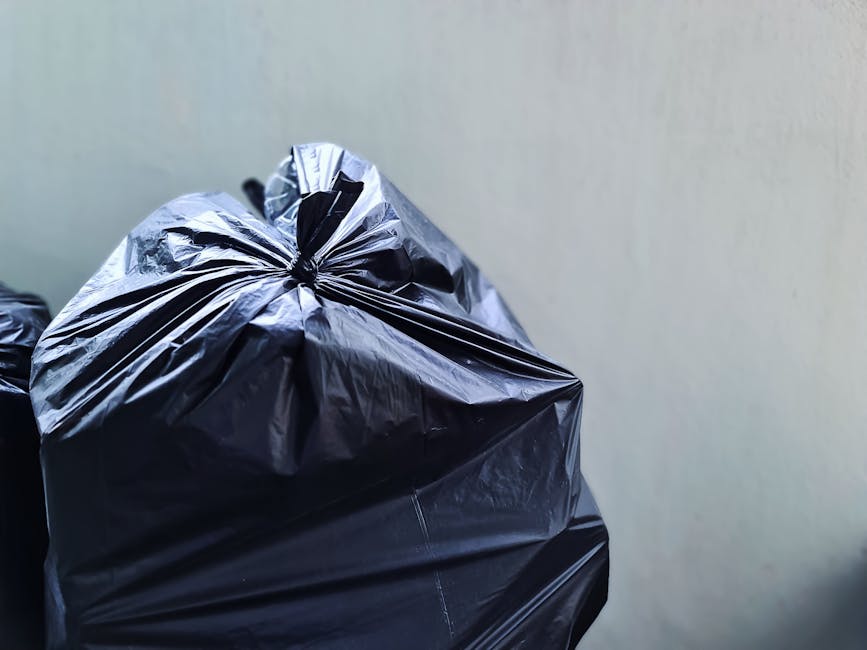 A close-up view of three large black rubbish bags made from shiny plastic, filled with waste materials and tightly knotted at the top, set against a neutral grey wall. The bags are positioned on a paved or concrete surface, possibly on a driveway or pavement area, with the leftmost bag partially overlapping the others. The environment appears to be outdoors or in a covered utility space, with natural lighting highlighting the glossy texture of the bags. These bags represent a typical method of private rubbish disposal, often used in independent collection services like those offered by Rubbish Removal Chelsea. The scene emphasizes the practical aspect of on-site waste management, with no visible tools or disposal equipment present. The background remains plain, ensuring the focus remains solely on the refuse bags, aligning naturally with themes of rubbish removal and alternative waste handling solutions.